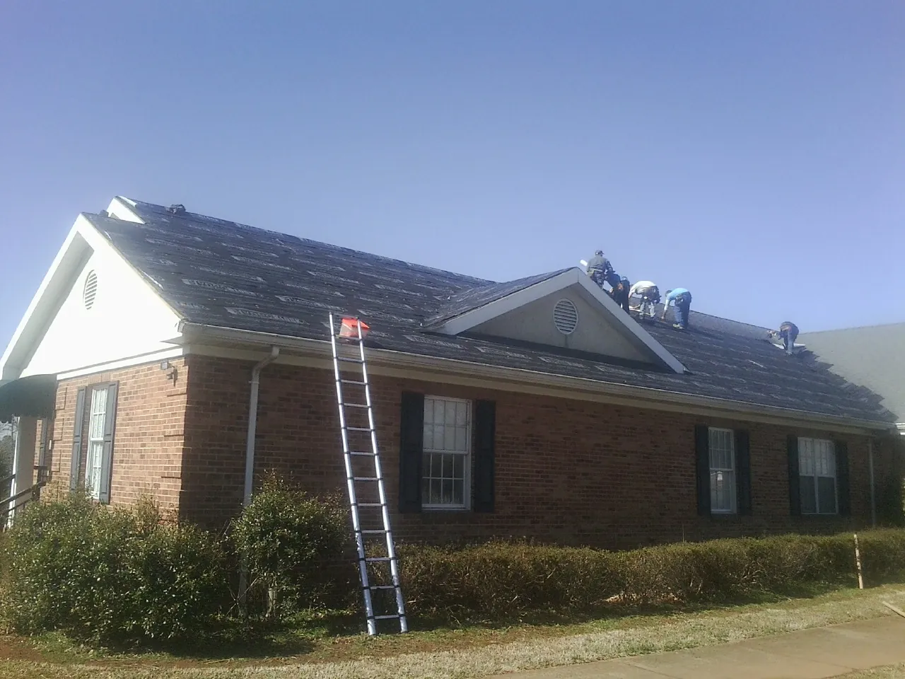 Crew tearing shingles off a brick ranch roof with a ladder set — Metal Roof Repair work by our crew in Burnt Cabins