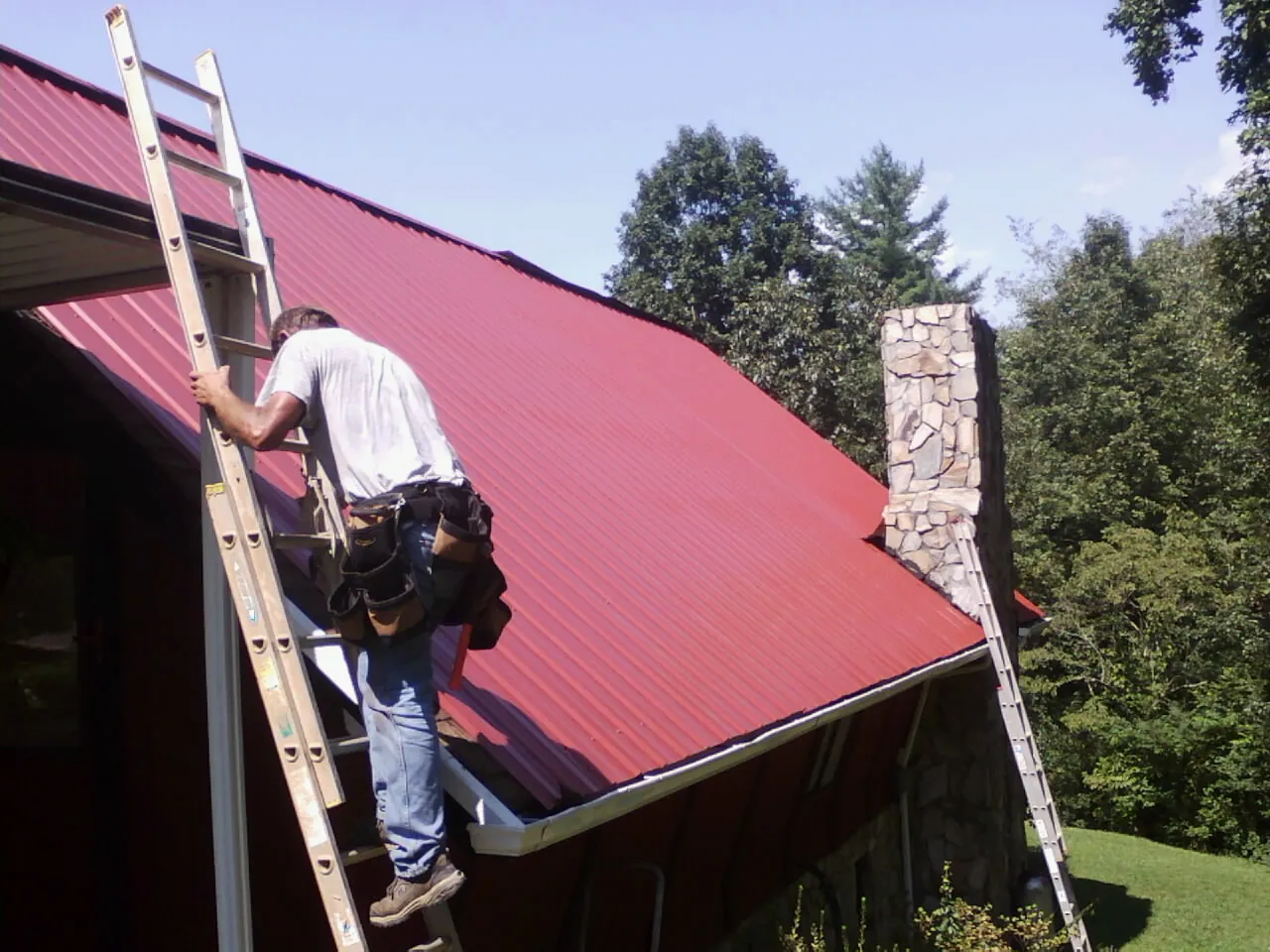 Roofer on a ladder beside a red metal roof and stone chimney — Soffit & Fascia Repair by Roofing Brothers in Burnt Cabins